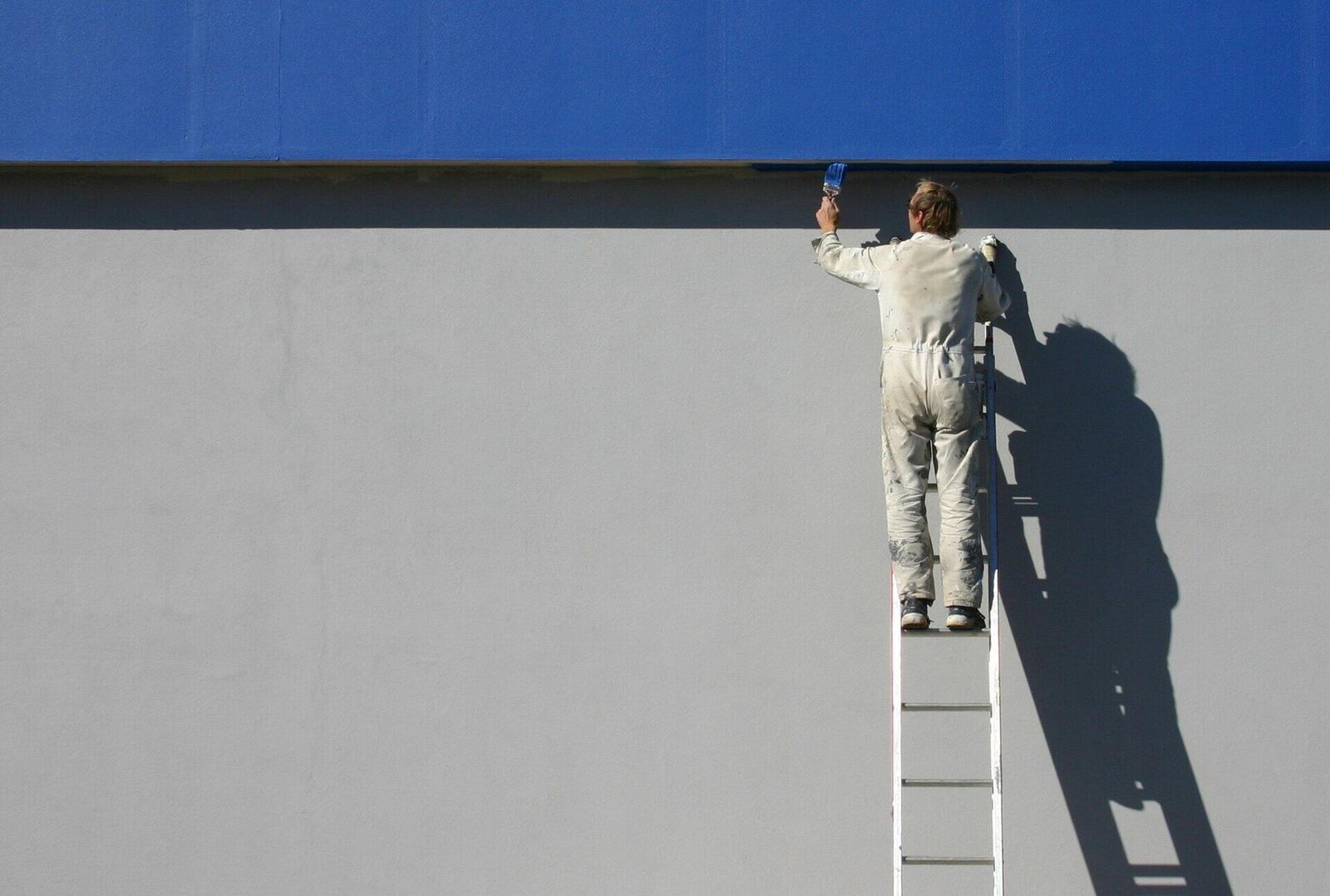 A painter in white overalls on a ladder rolling paint onto a wall
